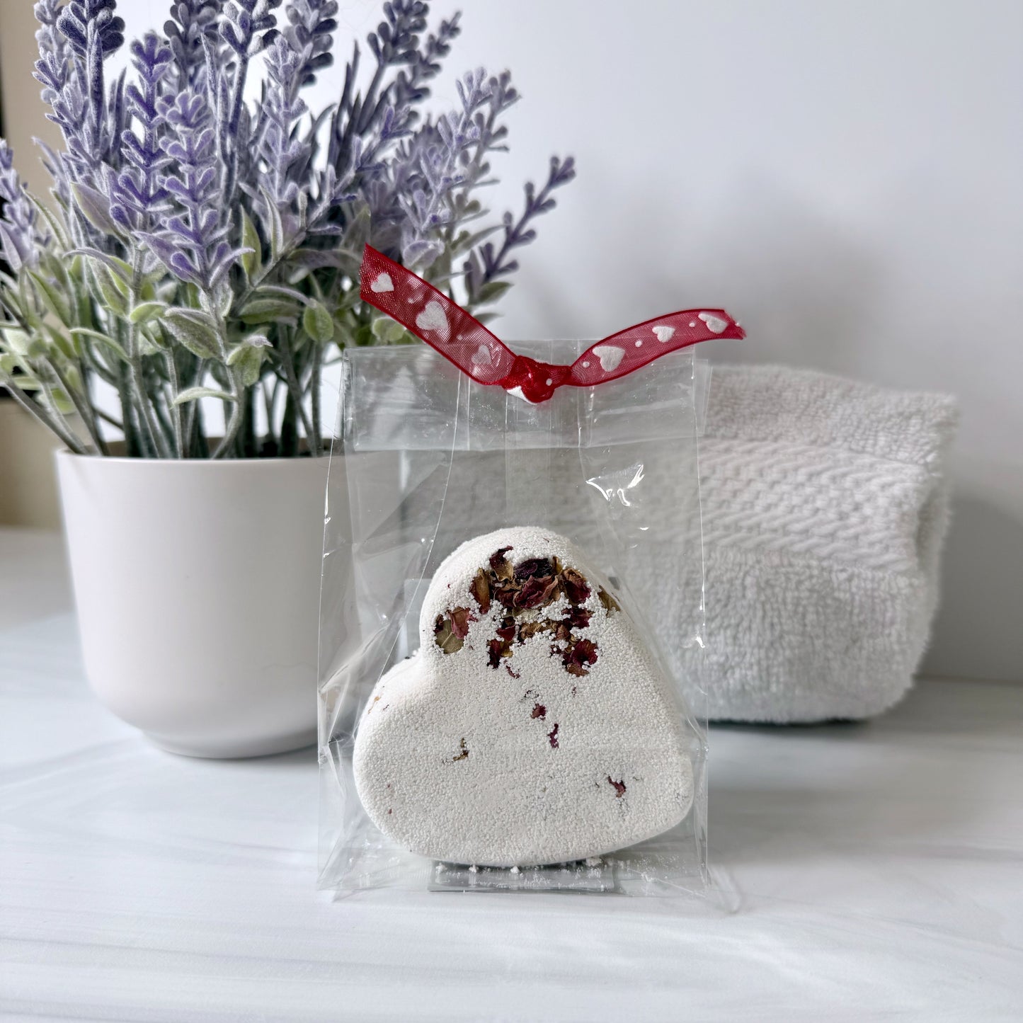 Heart-shaped bath bomb in clear packaging with a red ribbon, placed on a white surface with a plant in the background.